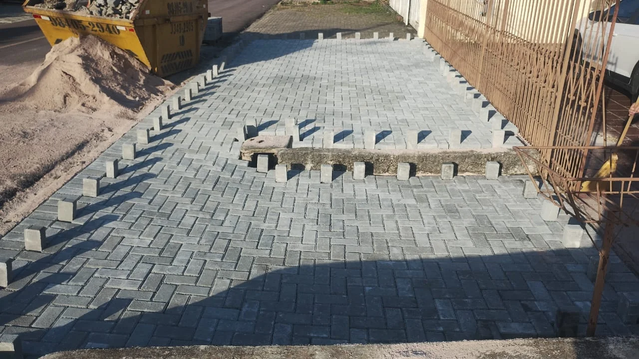 Assentamento em andamento, com peças soltas, areia e caçamba ao lado da entrada.